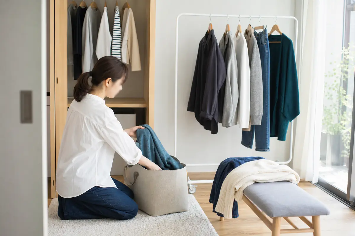 明るい室内で、女性が衣類ラックと開いたクローゼットの前に立ち、無地の服を仕分ける様子